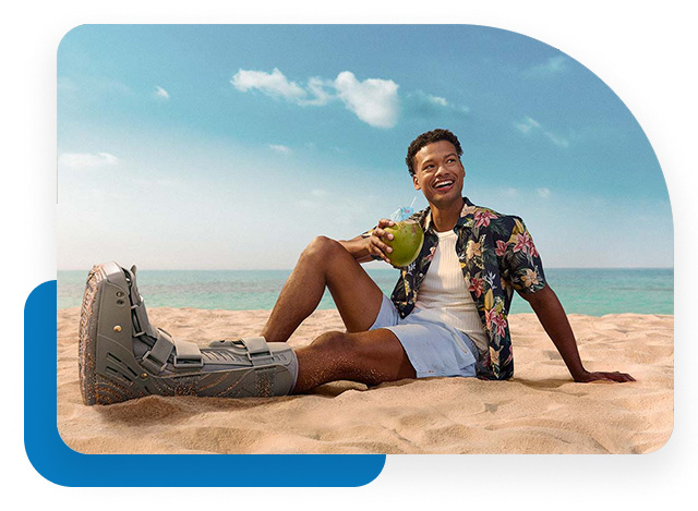 Young man sitting in the sand by the sea with his left foot in a cast.
