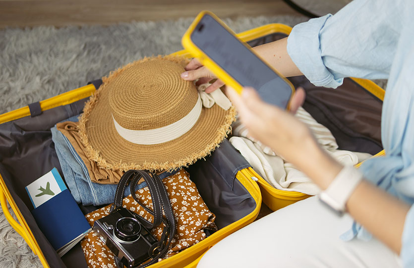 Woman carefully packing vacation essentials into a yellow suitcase, organizing travel documents, fashionable hat, camera, and clothing, preparing for an exciting trip.
