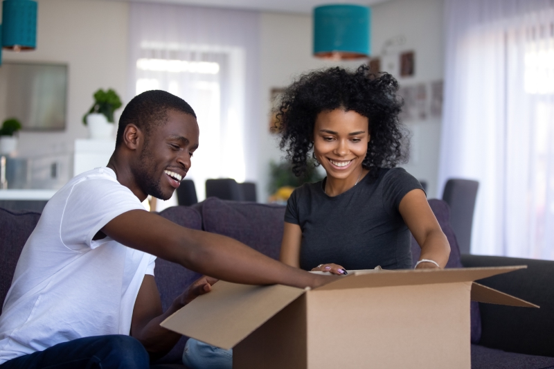 Young couple opening a cardboard box, sitting on the sofa in the living room, on moving day in Canada.