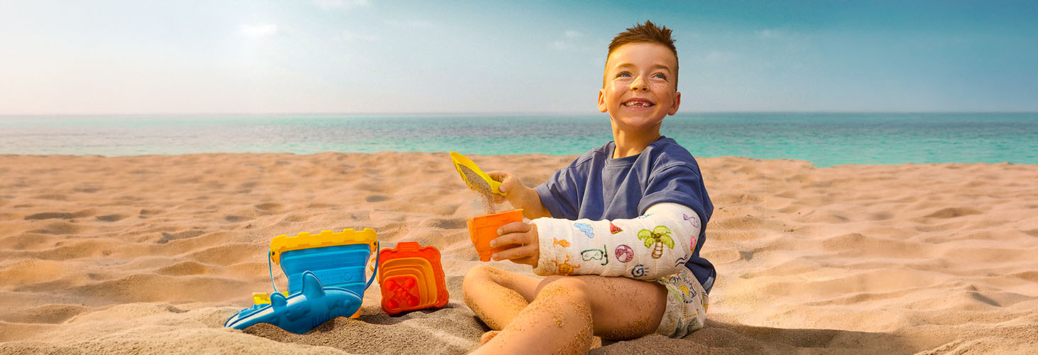A smiling little boy sitting in the sand and playing, with his arm in a cast.