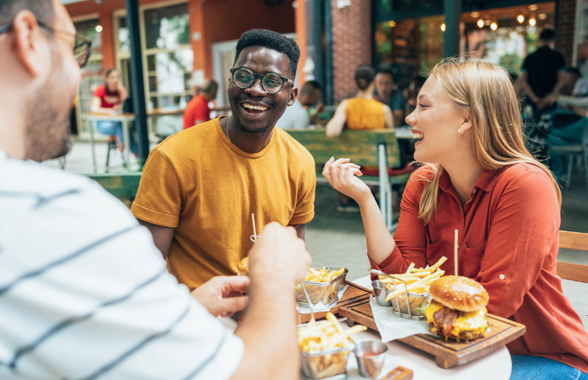 People sitting at an outdoor restaurant table, sharing a casual meal with burgers and fries on a sunny day.