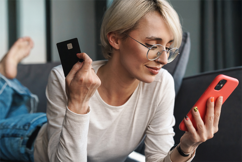 Person lying on a couch, holding a bank card in one hand and a mobile phone in the other, making an online purchase.