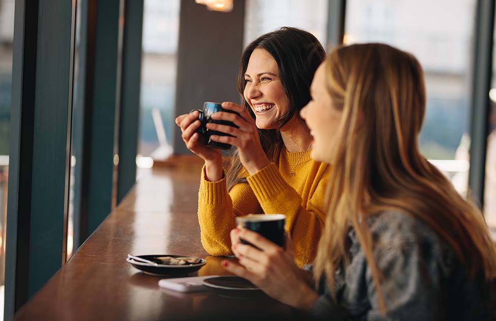 Two female friends smiling and sharing a coffee, happy to see each other again after their vacations.