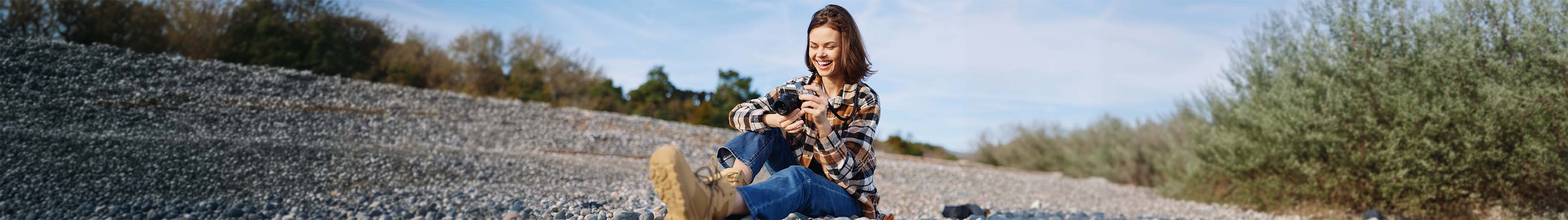 Person sitting on a pebble beach holding a camera, wearing a plaid shirt, jeans, and hiking boots, with trees and a clear sky in the background.