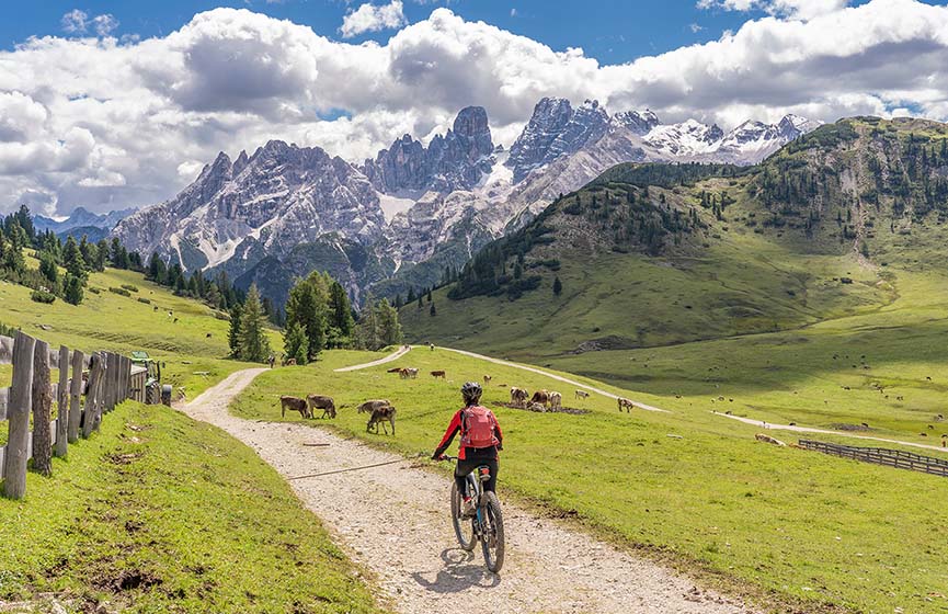 Active senior woman riding her electric mountain bike on the high plateau of Pratto Piazzo in the three peaks Dolomites , rocky silhouette of Mount Cristallo in background, South Tirol, Italy 