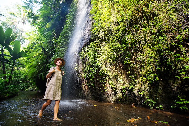 A person walking in a shallow stream beneath a waterfall, surrounded by lush vegetation
